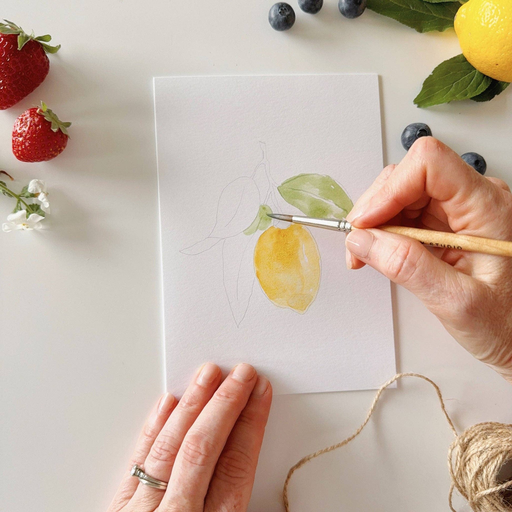 Person painting a lemon on a piece of paper with fruits and berries around