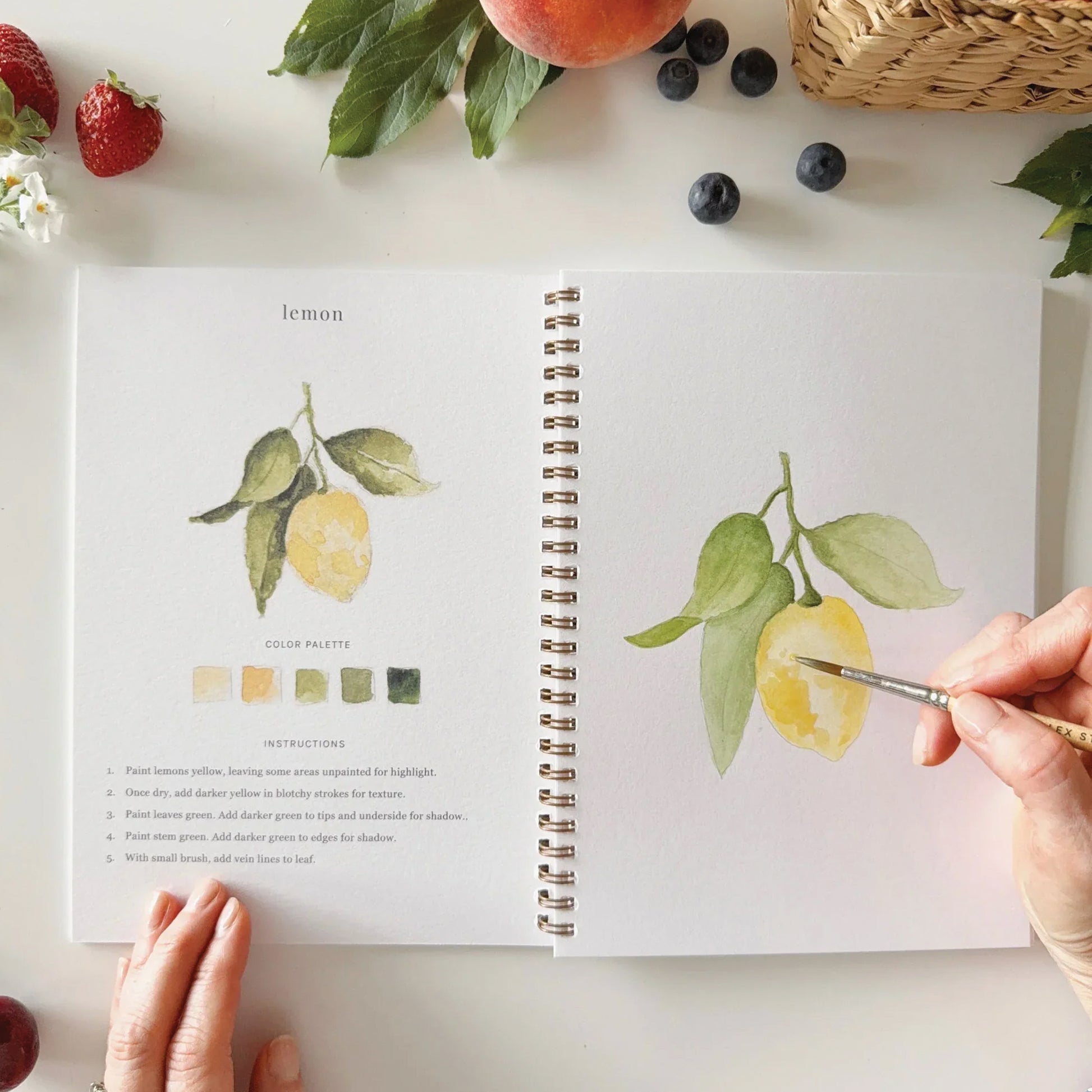 Open notebook with lemon illustrations and color palette on a table with fruits and a hand holding a paintbrush.
