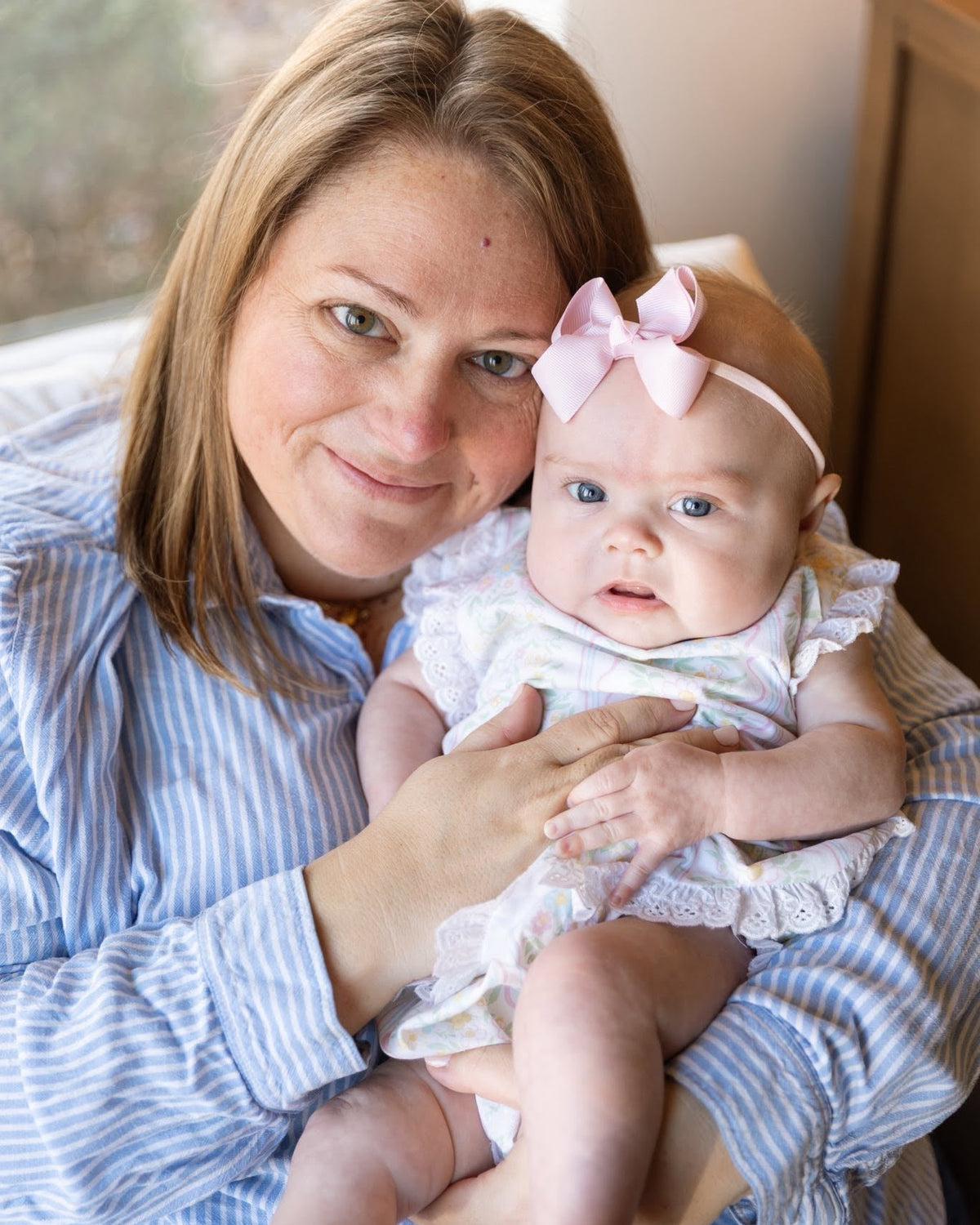 Woman holding a baby with a pink bow headband indoors.